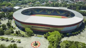 Vue aérienne d'un grand stade sportif entouré d'arbres lors d'une journée ensoleillée.