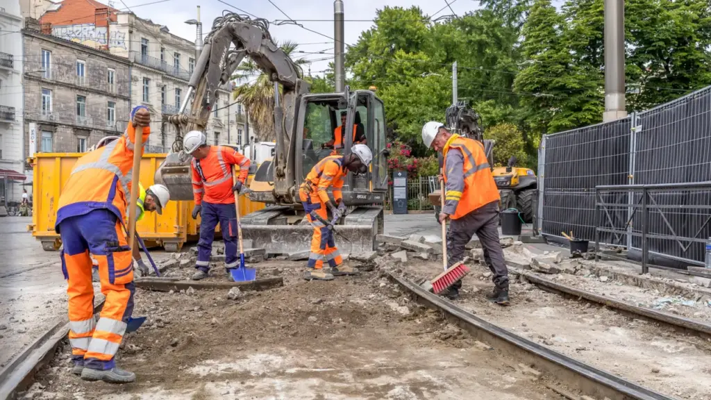 Ouvriers en gilets orange réparant une voie ferrée urbaine avec une pelle mécanique.