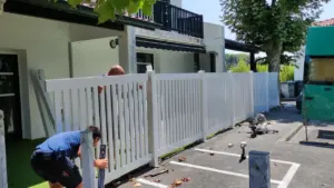 Deux ouvriers installent une nouvelle clôture en PVC blanc devant une maison résidentielle ensoleillée.