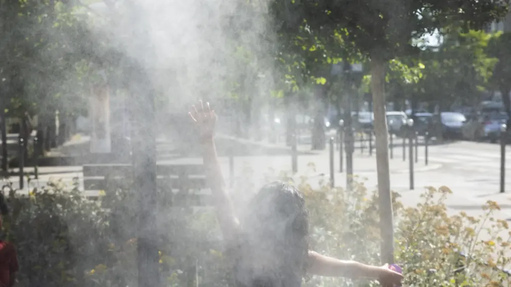 Une personne de dos lève les mains dans un brumisateur public en plein soleil.