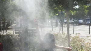 Une personne de dos lève les mains dans un brumisateur public en plein soleil.