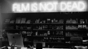Enseigne néon affichant 'Film is not dead' au-dessus de rayonnages de pellicules photo.