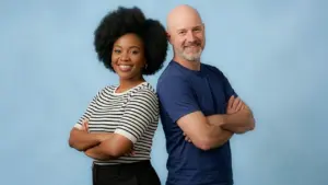 Une femme noire et un homme chauve sourient, posant bras croisés sur fond bleu clair.