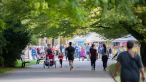 Des coureurs et des familles marchent sur une allée arborée vers des stands blancs.