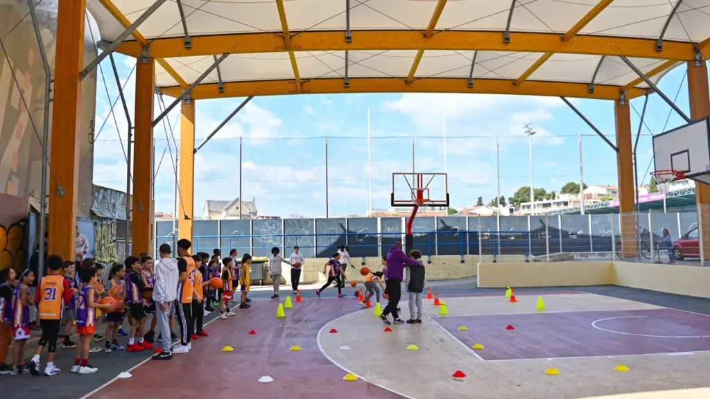 Groupe d'enfants s'entraînant au basketball sur un terrain extérieur couvert sous une structure moderne.