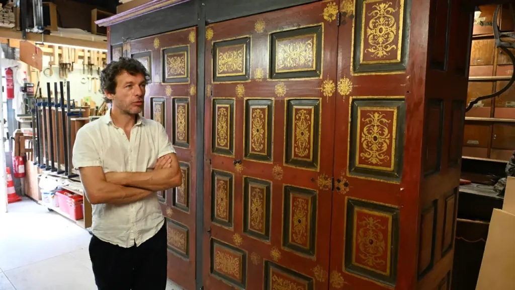 Un homme aux bras croisés debout devant une grande armoire ancienne en bois rouge décorée.